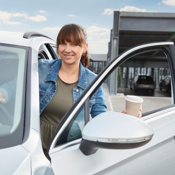 Frau steigt lächelnd aus Auto aus und hält Kaffeebecher in der Hand